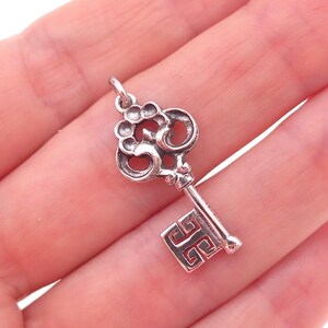 May include: A silver-toned metal key charm featuring an ornate, rounded bow with circular and scroll-like cutouts. The shaft is straight, leading to a rectangular bit decorated with a geometric, maze-like pattern and a small loop at the top.