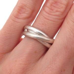 May include: A polished silver ring featuring a triple-band design where three smooth, rounded metal bands are intertwined to form a single cohesive piece of jewelry. The silver surface has a reflective, metallic finish with a continuous loop.