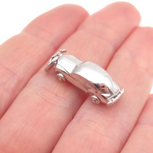 May include: A small, shiny silver car charm with detailed wheels and a classic car design. The charm is held in a person's hand, showcasing its size and reflective surface.