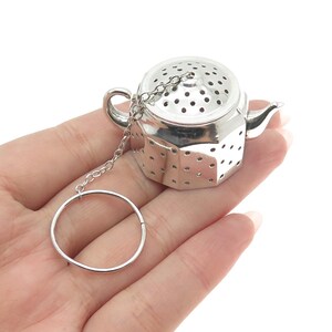 May include: Silver teapot-shaped tea infuser with a chain and a ring. The infuser has a mesh design with small holes.