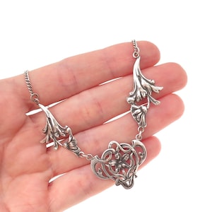 May include: A silver necklace featuring three ornate, floral-inspired metal links connected by a delicate chain. The central link displays a detailed flower motif, while the two side links showcase elongated, sweeping leaf-like patterns in polished silver.