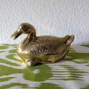 Könnte beinhalten: Eine goldfarbene Messing-Entenfigur mit detaillierten Federätzungen. Die Ente hat einen offenen Schnabel und sitzt auf einer grün-weiß gemusterten Oberfläche. Ein dekorativer Artikel.