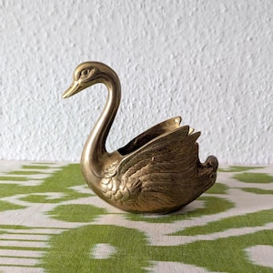 Puede incluir: Una figura de cisne de latón pulido con texturas de plumas detalladas a lo largo de las alas y el cuerpo. El cisne se representa con el cuello curvado elegantemente hacia adelante, y el centro hueco del cuerpo está abierto, mostrando una superficie interior lisa.