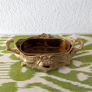 May include: Ornate gold-colored metal decorative bowl with handles. The bowl has an oval shape with a detailed, embossed design. The interior is a darker gold color. The bowl is sitting on a green and white patterned surface.