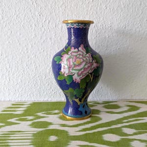Pode incluir: Um vaso azul cloisonné com um design floral. O vaso apresenta uma grande peónia rosa e branca com folhas verdes. A borda e a base são adornadas com ouro. O fundo é uma parede branca texturizada e uma superfície estampada em verde e branco.