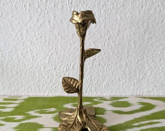 May include: A brass candlestick holder shaped like a single rose on a stem. The base consists of three flat, textured leaves that support the upright stem, which features two additional leaves and a rose-shaped top designed to hold a taper candle.