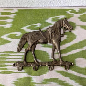 Op de afbeelding: Een zilverkleurige metalen wandhaak met een paard met zadel. Het paard is gemonteerd op een stang met drie haken. De achtergrond is een groen en wit patroon.