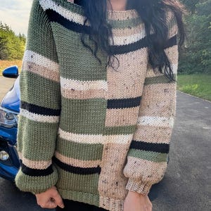 以下が含まれることがあります： パッチワークデザインのオーバーサイズセーター。オリーブグリーンとベージュのブロックが交互に配置され、黒、白、ベージュのストライプがアクセントになっています。袖口はリブ編みで、手編みのようです。