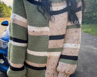 Könnte beinhalten: Ein übergroßer Patchwork-Pullover in Olivgrün und Beige. Das Design umfasst Farbblöcke mit schwarzen und weißen Streifen. Die Ärmel haben gerippte Bündchen. Der Pullover scheint handgestrickt zu sein.