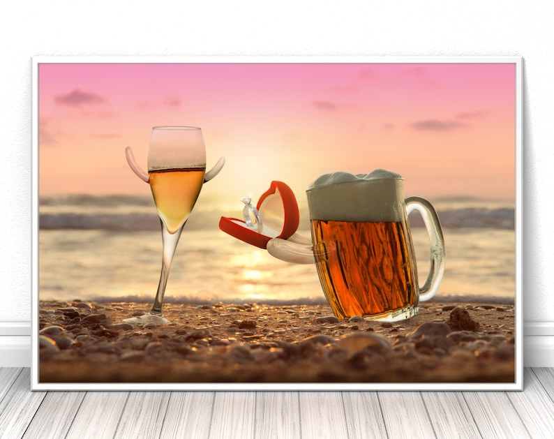 May include: A glass of white wine and a glass of beer on a beach at sunset. The beer glass is holding a red ring box with a diamond ring inside.