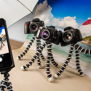 May include: Three cameras on flexible tripods, one with a red umbrella, are arranged on a beach scene backdrop. A smartphone on a tripod is taking a picture of the cameras.