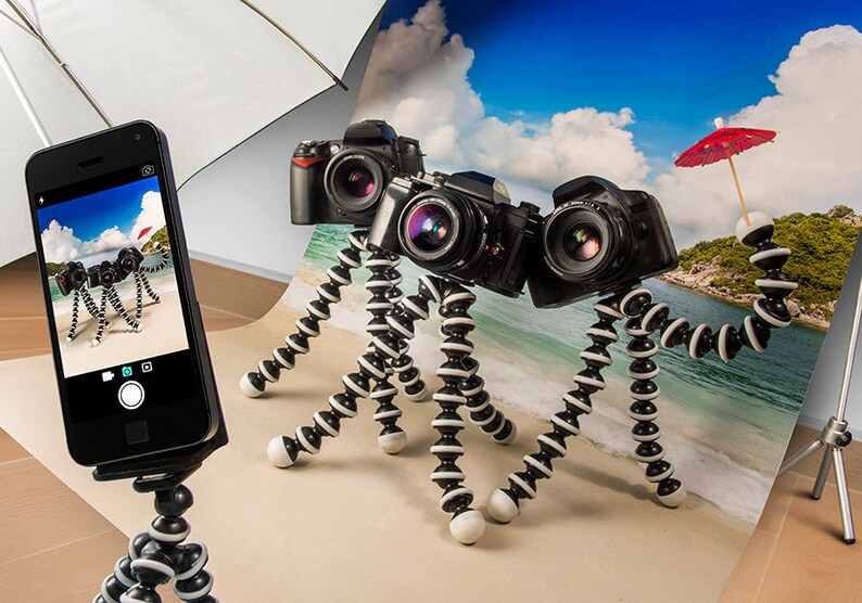 May include: Three cameras on flexible tripods, one with a red umbrella, are arranged on a beach scene backdrop. A smartphone on a tripod is taking a picture of the cameras.