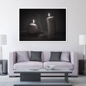 May include: A black and white photograph of a living room with a couch, two lamps, and a framed picture of two candles. The picture shows a white pillar candle and a round white candle, both lit.