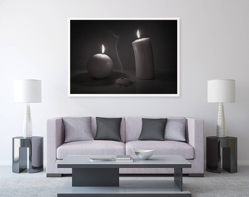 May include: A black and white photograph of a living room with a couch, two lamps, and a framed picture of two candles. The picture shows a white pillar candle and a round white candle, both lit.