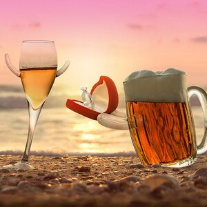 May include: A glass of champagne with a ring box in the shape of a hand proposes to a glass of beer on a beach at sunset.