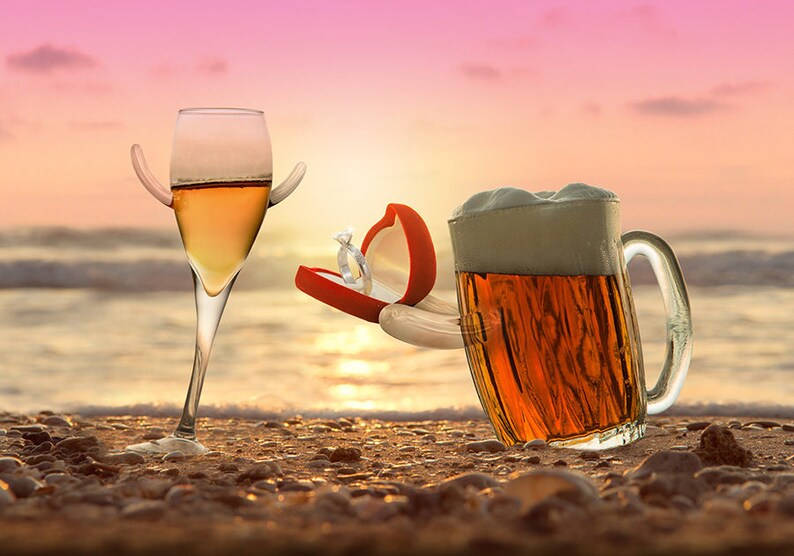 May include: A glass of champagne with a ring box in the shape of a hand proposes to a glass of beer on a beach at sunset.