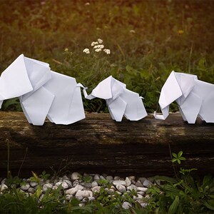May include: Three white origami elephants are lined up on a wooden log. The elephants are made of paper and are folded into a realistic shape. The elephants are all facing the same direction and are standing on a wooden log.
