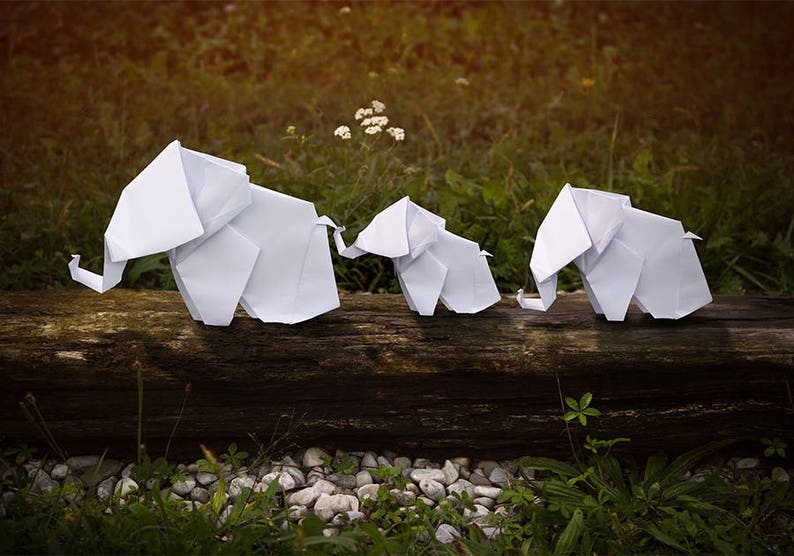May include: Three white origami elephants are lined up on a wooden log. The elephants are made of paper and are folded into a realistic shape. The elephants are all facing the same direction and are standing on a wooden log.