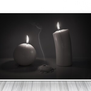 May include: Three white candles, two are lit and one is extinguished, with smoke rising from the extinguished candle. The candles are on a dark surface.