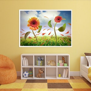 May include: A framed print of a colorful floral still life featuring three orange and red gerbera daisies with green leaves and petals blowing in the wind against a blue sky with white clouds. The flowers are set against a green field with orange and red petals scattered on the ground.
