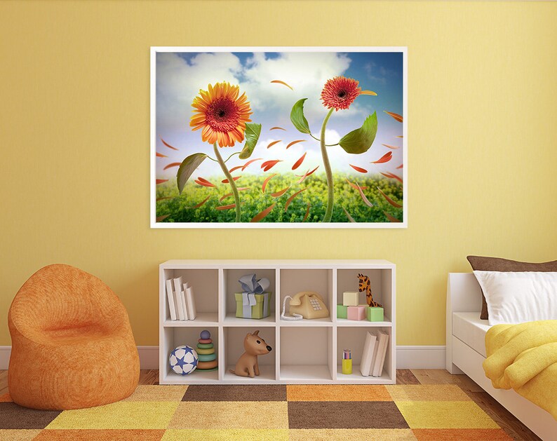 May include: A framed print of a colorful floral still life featuring three orange and red gerbera daisies with green leaves and petals blowing in the wind against a blue sky with white clouds. The flowers are set against a green field with orange and red petals scattered on the ground.