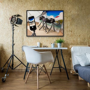 May include: A framed photo of a beach scene with a red umbrella, a smartphone, and three cameras on flexible tripods. The photo is hanging on a wall in a room with a desk, chair, and a couch.