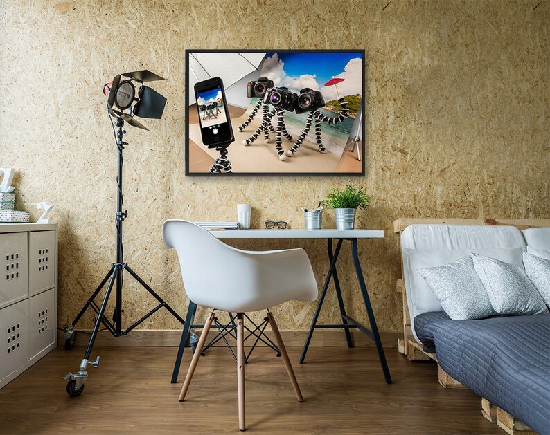 May include: A framed photo of a beach scene with a red umbrella, a smartphone, and three cameras on flexible tripods. The photo is hanging on a wall in a room with a desk, chair, and a couch.