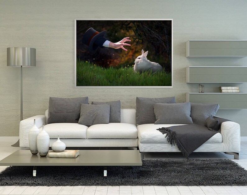 May include: A white rabbit sits in a grassy field, looking up at a hand reaching out from a top hat. The hand is wearing a black glove. The background is a blurred image of trees and bushes.