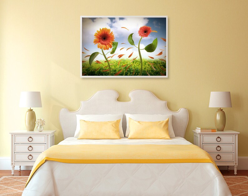 May include: A white bed with a yellow bedspread and two yellow pillows. A framed picture of two sunflowers with orange and red petals is hanging on the wall above the bed.