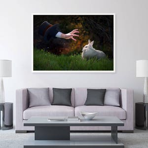 May include: A white rabbit sits in a grassy field, looking up at a hand reaching out from the darkness above. The hand is wearing a black top hat.