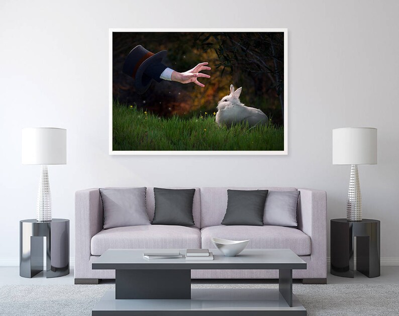 May include: A white rabbit sits in a grassy field, looking up at a hand reaching out from the darkness above. The hand is wearing a black top hat.