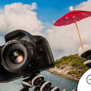 May include: A black DSLR camera with a lens attached is mounted on a tripod with a small red umbrella on top. The camera is pointed towards a tropical beach scene with blue water and green trees.