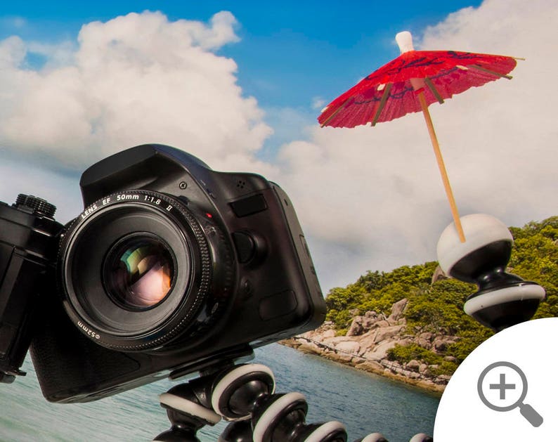 May include: A black DSLR camera with a lens attached is mounted on a tripod with a small red umbrella on top. The camera is pointed towards a tropical beach scene with blue water and green trees.