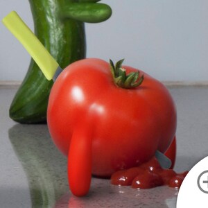 May include: A red tomato with a green stem is pierced by a yellow knife. The tomato is lying on a white surface with a red sauce stain.
