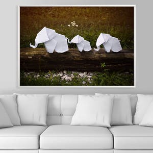 May include: Three white origami elephants are arranged on a log in a forest setting. The elephants are made of paper and are facing the same direction. The background is a blurred image of green foliage and brown earth.