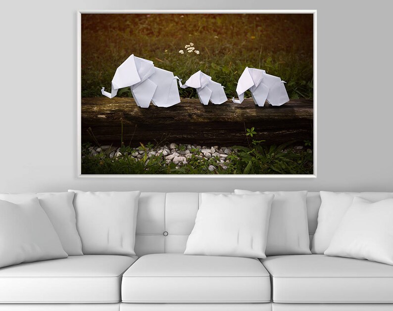 May include: Three white origami elephants are arranged on a log in a forest setting. The elephants are made of paper and are facing the same direction. The background is a blurred image of green foliage and brown earth.