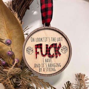 May include: Wooden Christmas ornament with a red and black plaid design. The ornament says "OH LOOK! IT'S THE LAST FUCK I HAVE. AND IT'S HANGING BY A STRING."