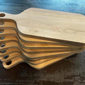 May include: A stack of eight light-coloured wooden cutting boards, each featuring a rectangular body with rounded corners and a handle with a circular hole at the end. The boards are arranged in a fanned-out pattern, showcasing the natural wood grain.