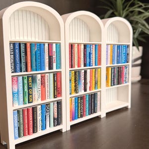 May include: Two white miniature arched bookshelves filled with tiny colourful books. Each shelf features rows of miniature volumes with legible titles like Hello Beautiful, The Midnight Feast, Beach Read, and Mad Mabel, arranged in a vertical display.