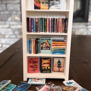 May include: A white arched miniature bookshelf with four shelves filled with tiny book replicas. The shelves contain various miniature books arranged upright and stacked, while additional miniature books are scattered on the surface below.