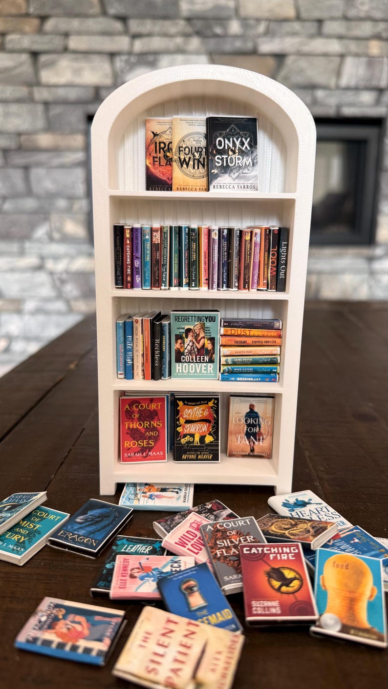 May include: A white arched miniature bookshelf with four shelves filled with tiny book replicas. The shelves contain various miniature books arranged upright and stacked, while additional miniature books are scattered on the surface below.