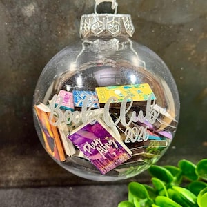 May include: A clear spherical glass bauble filled with miniature books, featuring a silver metal cap and a white ribbon hanger. The exterior is etched with the white text Book Club 2026. The tiny books inside have colourful covers.