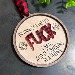 May include: Round wooden ornament with a red and black plaid design. The ornament says "Oh look! It's the last F**k I have. And it's hanging by a string."