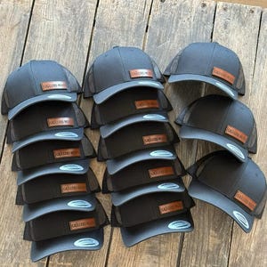 Può includere: Una pila di cappellini da camionista grigio scuro con retro in rete nera e visiere grigio chiaro. Ogni cappellino presenta una toppa in pelle marrone con la scritta "GRILLERS MEAT". I cappellini sono disposti su una superficie di legno.