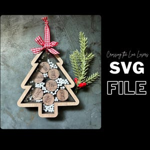 May include: A wooden Christmas tree ornament with a red and white checkered bow. The ornament has a clear plastic insert that holds small white beads and round wooden discs with names written on them. The text "Crossing the Line Lasers SVG FILE" is visible in the image.