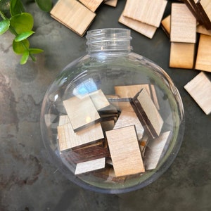 May include: A clear, spherical plastic ornament filled with small, rectangular wooden tiles. The tiles have light-colored faces and dark-colored edges. Additional loose wooden tiles are scattered around the base of the ornament.
