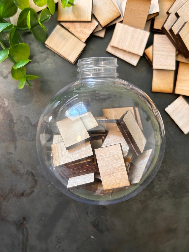 May include: A clear, spherical plastic ornament filled with small, rectangular wooden tiles. The tiles have light-colored faces and dark-colored edges. Additional loose wooden tiles are scattered around the base of the ornament.