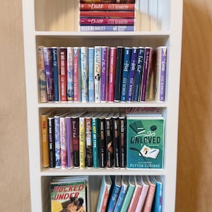 May include: A white arched miniature bookshelf filled with rows of small books. The shelves contain various titles including The Dare, The Chase, Unloved by Peyton Corinne, and Pucked Under by Helena Hunting, arranged in upright and stacked positions.