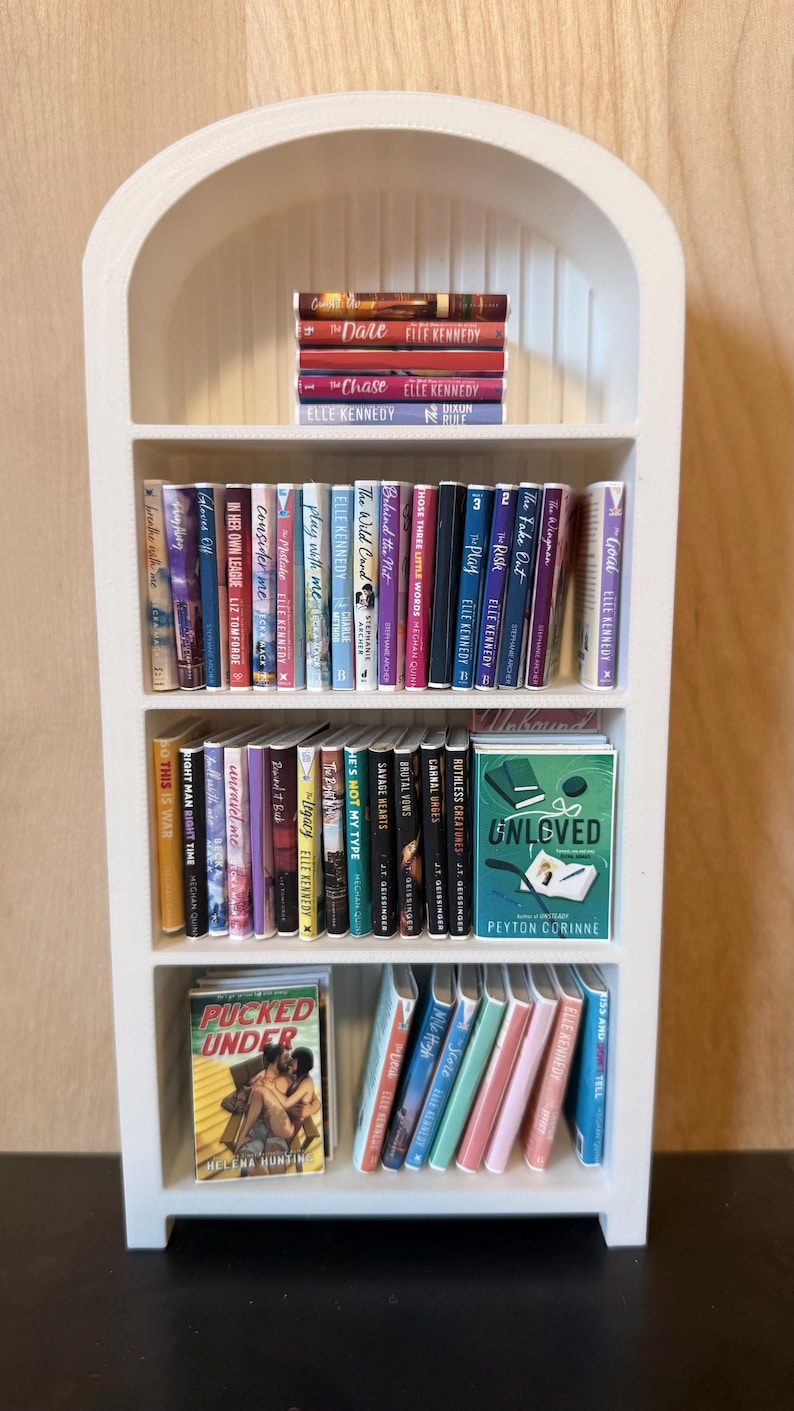 May include: A white arched miniature bookshelf filled with rows of small books. The shelves contain various titles including The Dare, The Chase, Unloved by Peyton Corinne, and Pucked Under by Helena Hunting, arranged in upright and stacked positions.