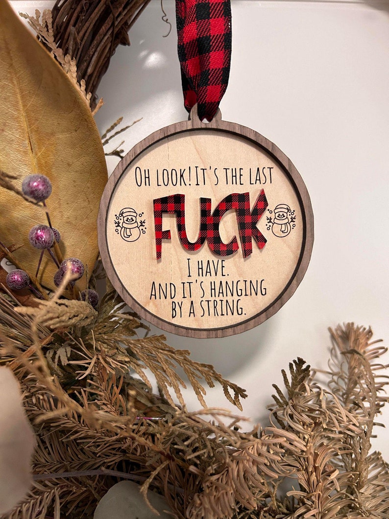 May include: Wooden Christmas ornament with a red and black plaid design. The ornament says "OH LOOK! IT'S THE LAST FUCK I HAVE. AND IT'S HANGING BY A STRING."